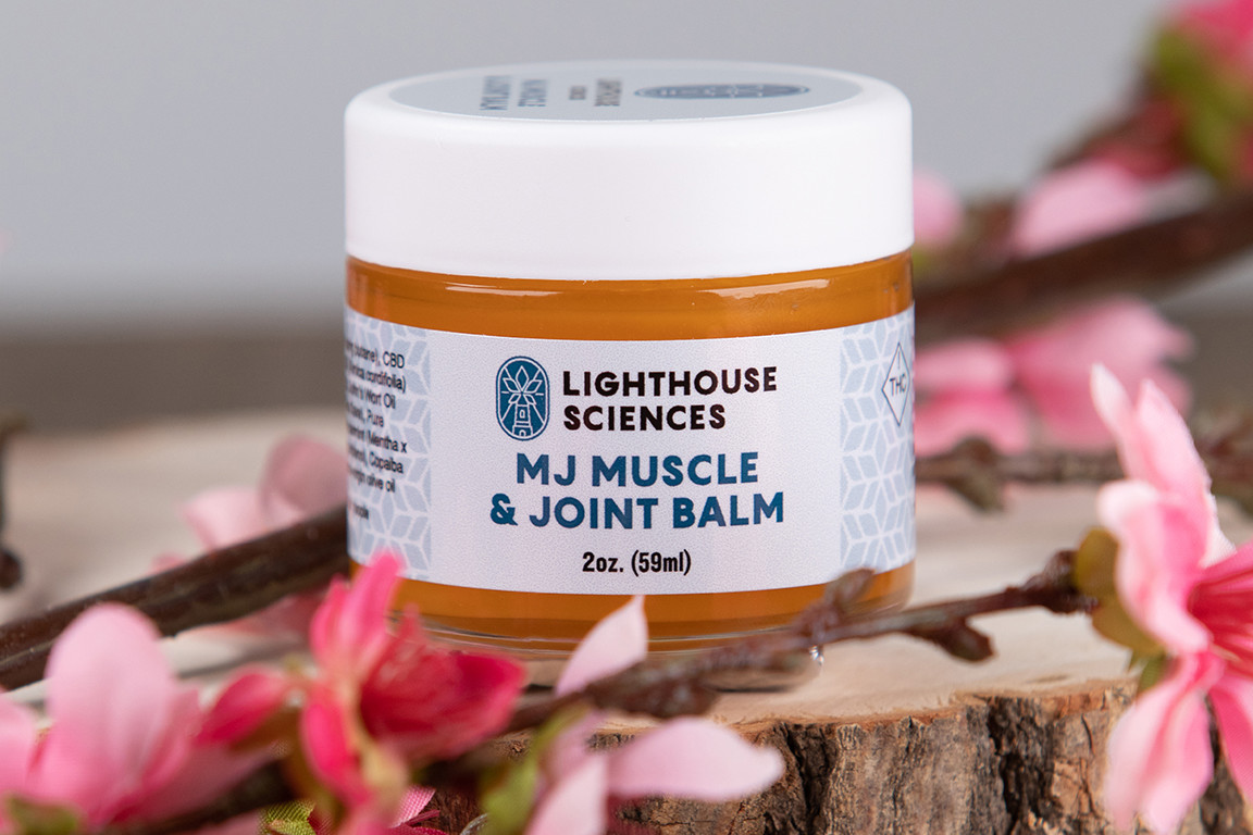 Lighthouse Sciences MJ Muscle & Joint Balm on a wood stop surrounded by flowers
